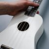 A hand strumming a white ukulele with focus on the strings and finger placement.